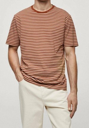 Uomo che indossa una maglietta a maniche corte a righe orizzontali arancione ruggine e bianca con pantaloni beige chiaro, una mano in tasca, su sfondo uniforme.