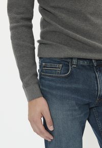 Bărbat îmbrăcat într-un pulover tricotat gri și blugi blue denim, având mâna stângă relaxată lângă corp, pe un fundal simplu.
