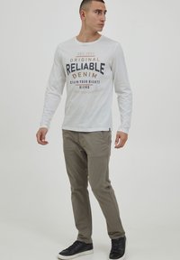 Langermet hvit t-skjorte med navy og oransje tekstdesign. Lys grå bukser, sorte joggesko. Enkel, uformell stil med rund hals.