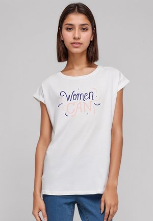 Young woman with straight brown hair wears a white T-shirt with "Women Can" text in purple and pink, paired with blue jeans.