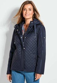 Marineblaue, gesteppte Jacke mit Kapuze, ausgestattet mit Druckknöpfen, Seitentaschen und elastischen Bündchen, kombiniert mit einem gestreiften Hemd und blauen Jeans.