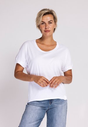 Woman with short blonde hair wearing a white rolled-sleeve t-shirt and blue jeans, standing with hands gently clasped.
