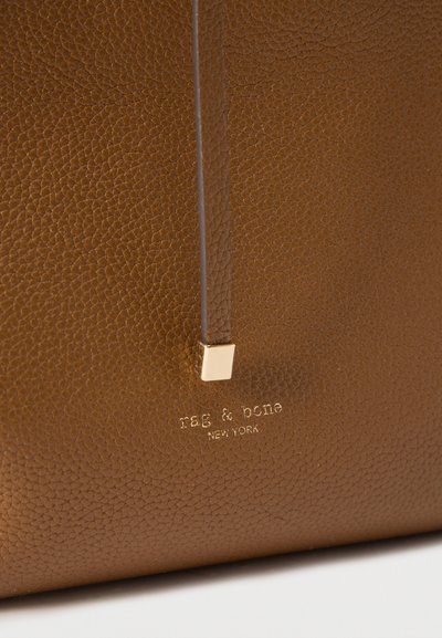 Κοντινό πλάνο καφέ υφής δέρματος με το ανάγλυφο λογότυπο «rag & bone New York» και ένα δερμάτινο λουράκι που τελειώνει σε μια μικρή χρυσή τετράγωνη λεπτομέρεια.