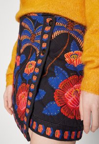 Femme portant une jupe noire matelassée avec un imprimé floral bleu et rouge et un devant boutonné, assortie à un pull jaune en laine pelucheuse.