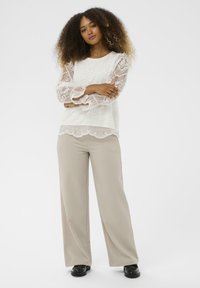 Blouse en dentelle blanche avec des motifs floraux raffinés et des manches à volants, associée à un pantalon beige ample et des chaussures plates noires.