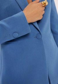 Niebieska marynarka wykonana z teksturowanej tkaniny, z wyłogowym kołnierzem, dwoma okrągłymi guzikami na mankietach oraz jedną kieszonką na piersi.