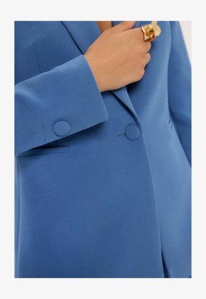 Niebieska marynarka wykonana z teksturowanej tkaniny, z wyłogowym kołnierzem, dwoma okrągłymi guzikami na mankietach oraz jedną kieszonką na piersi.