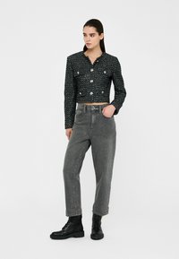 Giacca nera textured cropped con grandi bottoni abbinata a jeans grigi a vita alta e stivaletti neri con lacci. Sfondo neutro.