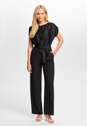 Femme blonde portant un haut en satin noir et un pantalon noir à jambes larges avec une ceinture nouée à la taille, posant avec une main dans la poche et des talons aux pieds.