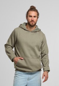 Sudadera color verde oliva con un bolsillo frontal, capucha con cordón y mangas largas. Combinada con unos jeans de mezclilla azul claro. Material suave y casual.