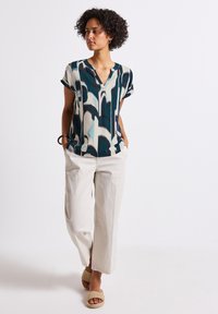 A woman stands confidently, wearing a patterned top and light-colored trousers, with hands in pockets, against a plain background.