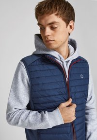 Marine isoleret vest med horisontalt quiltet struktur, rød lynlås og grå hætte under. Har et logo på venstre bryst.