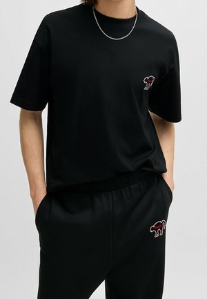 Person trägt schwarzes kurzärmeliges Shirt und Hose mit passendem Katzenlogo in Rot und Weiß, Hände in den Taschen, vor einfarbigem Hintergrund.