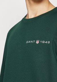 Mörkgrön bomullströja med rundad halsringning. Har vit broderad text "GANT 1949" och en sköldlogotyp på vänster bröst.