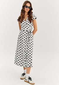 Černobílé puntíkované zavinovací šaty s krátkými rukávy, páskem v oblasti pasu a středně dlouhým lemem, doplněné sportovními bílými teniskami.