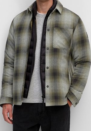 Mann trägt geschichtete Kleidung mit einer grünen und grauen karierten Hemdjacke über einer schwarzen gesteppte Weste und einem hellgrauen Strickpullover.