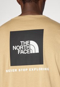 Μπεζ βαμβακερό T-shirt με μεγάλο μαύρο τετράγωνο που περιέχει λευκό κείμενο "THE NORTH FACE" και σλόγκαν "NEVER STOP EXPLORING" από κάτω.