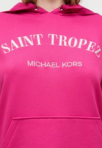 Sweat à capuche rose avec texte blanc brodé "SAINT TROPEZ" courbé au-dessus de "MICHAEL KORS" et une poche kangourou à l'avant visible.
