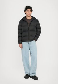Schwarze Pufferjacke mit Reißverschluss, Kapuze und gepolsterter Textur, kombiniert mit hellblauen, lockeren Jeans und schwarzen Schuhen.