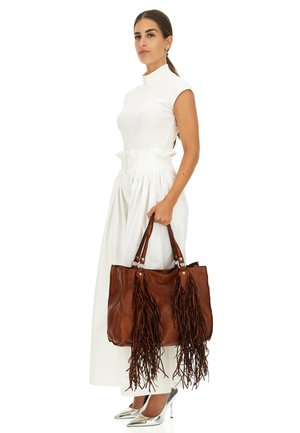 Borsa a tracolla in pelle marrone con frange, dettagli in argento e doppi manici. Modella indossa un top bianco aderente e una gonna lunga, abbinati a tacchi argento.