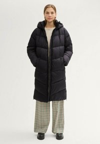 Cappotto imbottito nero con cappuccio, chiusura zip, cuciture verticali e tasche laterali, abbinato a pantaloni a gamba larga a quadri e scarpe nere.