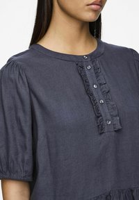 Blouse bleu foncé avec des manches courtes bouffantes, un plastron froncé avec des boutons et un ourlet légèrement froncé pour une silhouette décontractée.