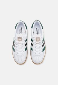 Scarpe da ginnastica bianche con strisce verdi e nere, tomaia in pelle texturizzata e rete, lacci piatti e suola in gomma. Dettaglio del logo in ottone sulla linguetta.