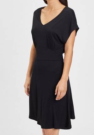 Vrouw draagt een zwart, kort mouw, V-hals jurkje met een getailleerde taille en een uitlopende rok, staand tegen een effen achtergrond.