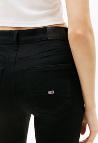 Person wearing black jeans with a small flag logo on the back pocket and a white cropped top, shown from the waist down.