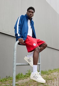 Veste de sport bleue avec des rayures blanches, short rouge arborant un logo, et baskets beiges avec des accents gris. Modèle assis sur un garde-corps en métal.