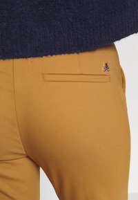 Slimfit byxor i senapsfärg med en slät yta, med en bakficka och en diskret logotypdetalj. Mörk tröja synlig ovanför.