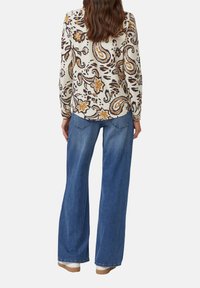 Vrouw in blauwe wijde jeans en een lange beige top met bruine en oranje bloemen paisleyprints, staand met haar rug naar ons gekeerd.