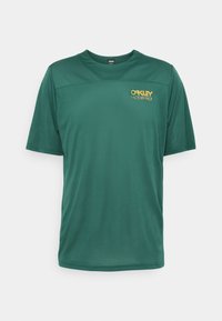 Zelené tričko s krátkými rukávy z lehké látky s kulatým výstřihem. Na hrudi jsou žluté loga „OAKLEY“ a „FACTORY PILOT“.