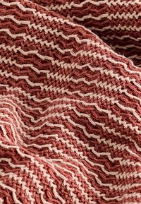 Couverture tissée texturée en terre cuite avec un motif en zigzag blanc cassé, présentant un design doux et côtelé ainsi que des bords effilochés visibles.