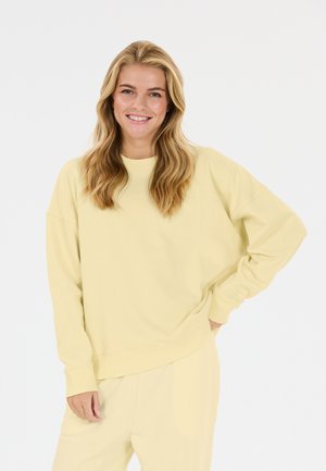 Femme souriante aux longs cheveux blonds portant un sweat-shirt jaune clair et un pantalon assorti, debout, une main sur la hanche.