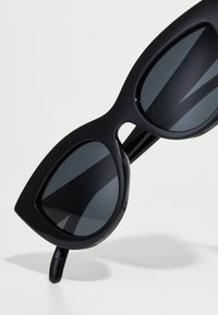 Mustat aurinkolasit, joissa on suuret, kulmikkaat linssit, paksu kehys ja tyylikäs, matta viimeistely. Linssit ovat tummat ja hieman heijastavat.