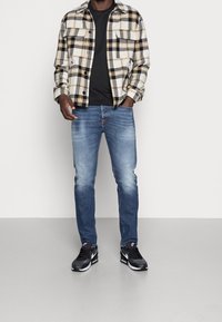 Rutigt flanellskjorta i krämfärg, svart och gult, kombinerad med blåjeans och svarta sportskor. Har två bröstfickor och knäppning framtill.