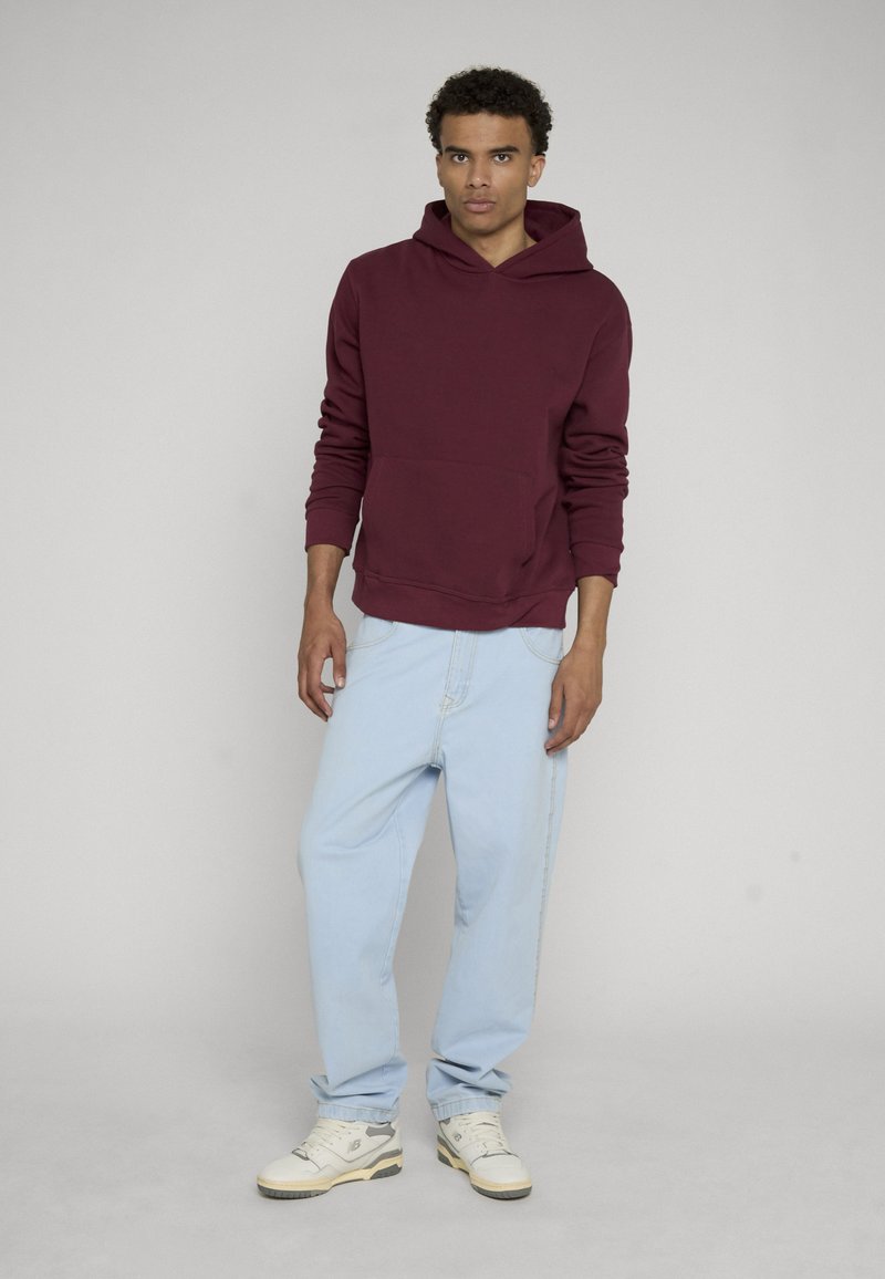 Jeune homme debout, portant un sweat à capuche bordeaux, un jean bleu clair et des baskets blanches, devant un fond uni gris clair.
