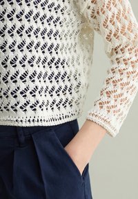 Maglione in pizzo color crema con maniche a tre quarti, caratterizzato da un motivo a diamante, abbinato a pantaloni blu navy e una mano in tasca.