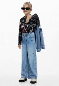 Černý top s dlouhým rukávem s bílým květinovým motivem a flitrovými doplňky, v kombinaci se svítvě modrými širokými džíny s vyšívanými detaily. Černé sluneční brýle.