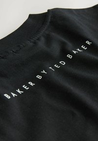 Melns audums ar apaļu kakla izgriezumu, kurā uzraksts "BAKER BY TED BAKER" baltā krāsā novietots horizontāli gar šuves līniju ap kaklu.
