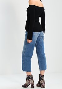 Mujer vistiendo un top negro de hombros descubiertos, jeans cortos de pierna ancha azul y botas de tobillo con estampado floral, de pie contra un fondo blanco.