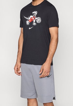 Homme portant un t-shirt noir avec un dinosaure cartoon jouant au basket-ball et un short gris Nike avec un logo noir.