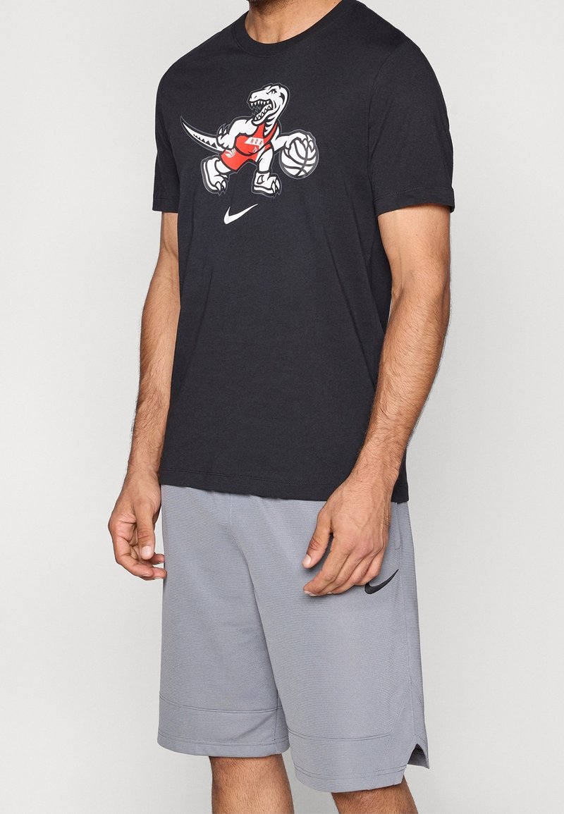 Homme portant un t-shirt noir avec un dinosaure cartoon jouant au basket-ball et un short gris Nike avec un logo noir.