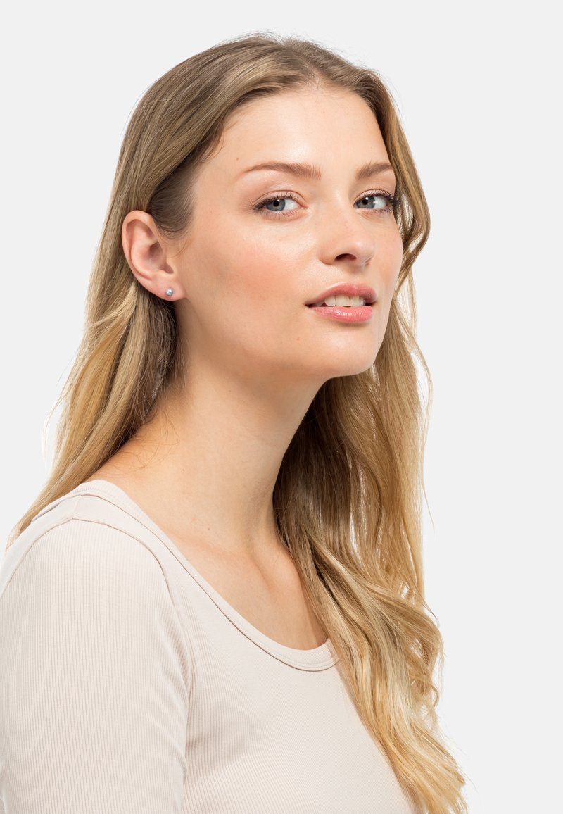 Orecchini a bottone con un semplice design rotondo in argento, caratterizzati da una pietra di colore azzurro chiaro. Il modello ha i capelli lunghi biondi e indossa un top neutro.