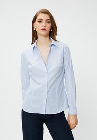 Camicia azzurra chiara con righe bianche verticali, colletto con punte, maniche lunghe e orlo curvo. Tessuto morbido e design aderente.