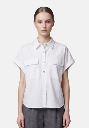 Giovane donna con capelli pettinati all'indietro, indossa una camicia bianca a maniche corte con bottoni e pantaloni grigi, in piedi su uno sfondo semplice.