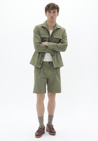 Olivgrünes Jacken- und Shorts-Set mit Knopfdesign, vorderen Taschen und strukturiertem Stoff. Braune Schuhe mit Schnürsenkeln und gerippten Socken.
