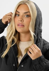 Svart huva jacka, med dragkedja och elastiska ärmslut. Stylat med en krämfärgad stickad tröja under, vilket framhäver texturkontrasten.