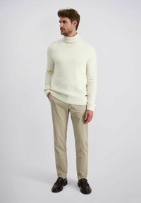 Man in een witte coltrui, beige broek en donkerbruine instappers, staand met handen in de zakken tegen een effen achtergrond.